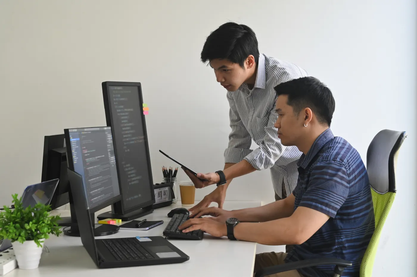 Two colleagues review records on a desktop screen. One sits at the keyboard. The other stands with a tablet.