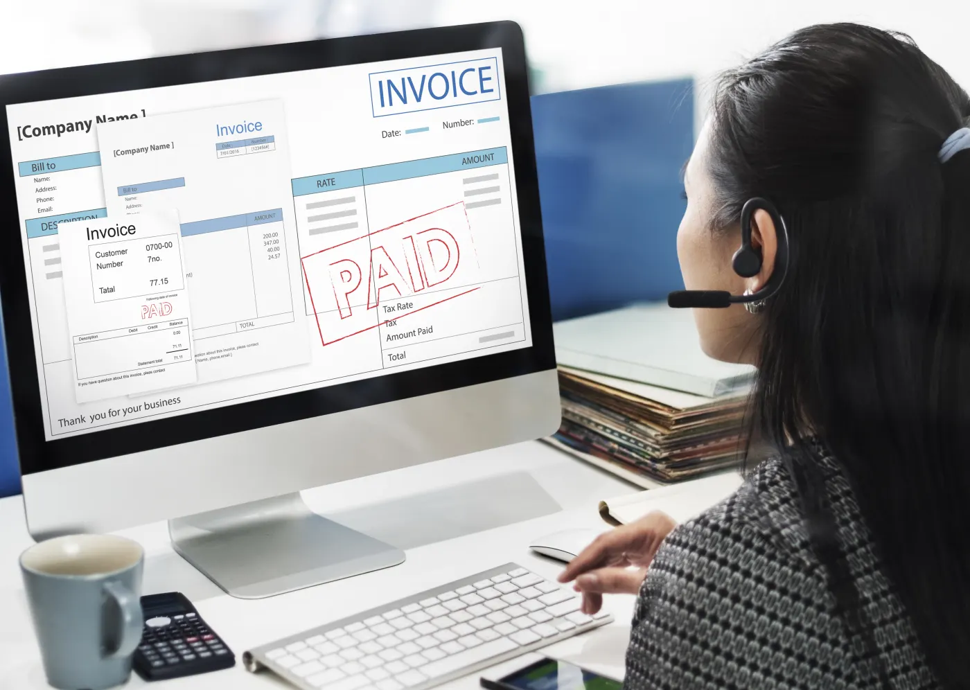 A person with a headset works at a desk. The computer screen shows invoices, and one invoice has a red PAID stamp.