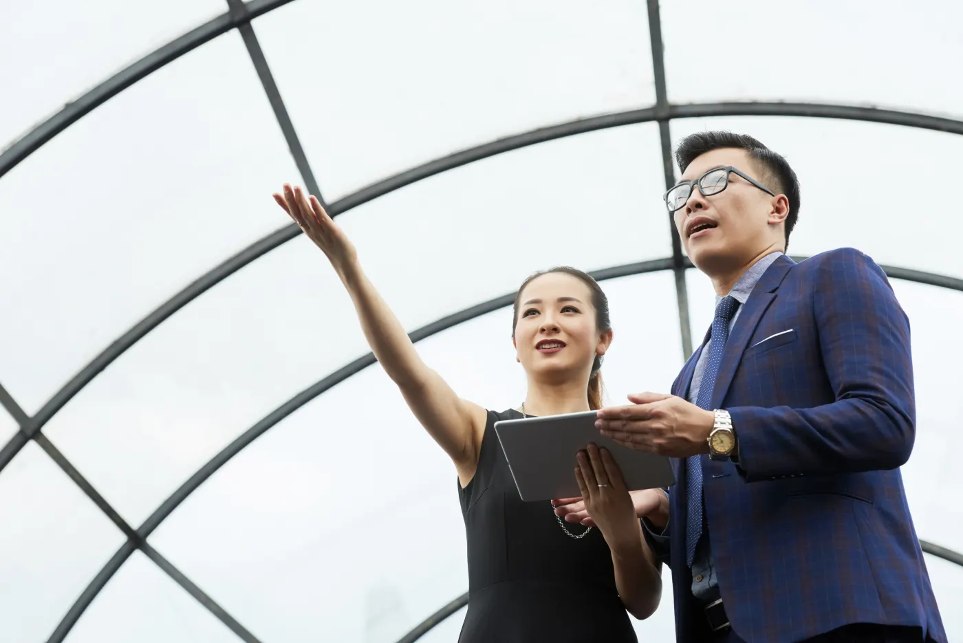 Two colleagues stand under a glass dome. One points upward. The other holds a tablet.
