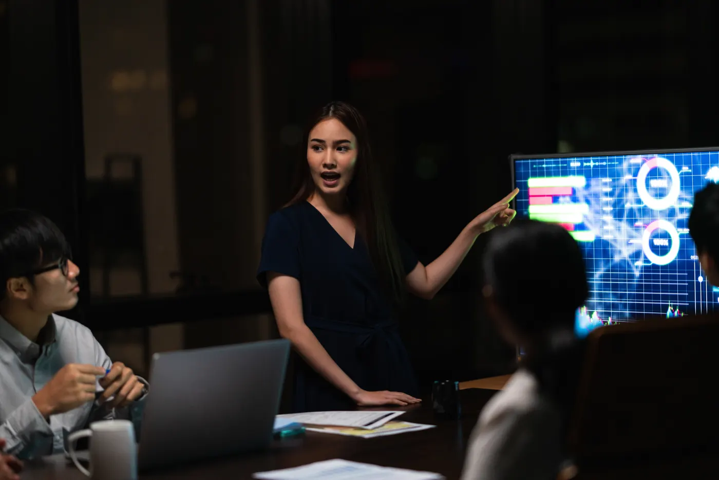 A presenter points at a wall screen with charts during an evening meeting. Colleagues sit at the table with laptops.